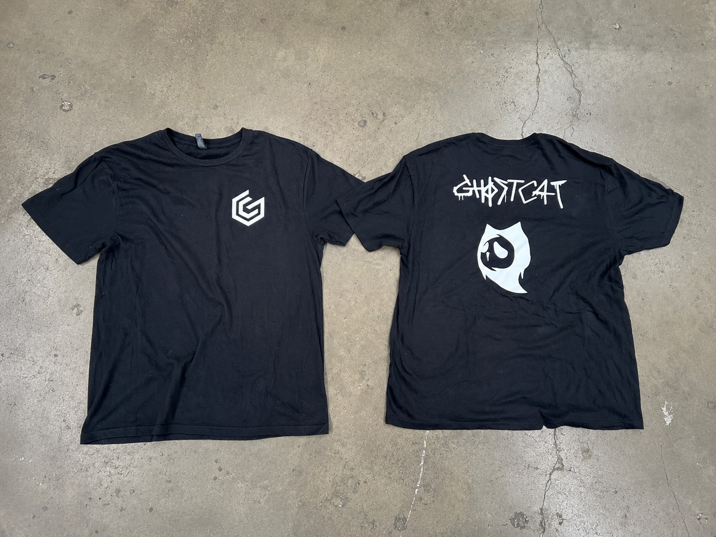 The original black GhostCat t-shirt laid flat on concrete, front and back, with the chest mark and the back graphic